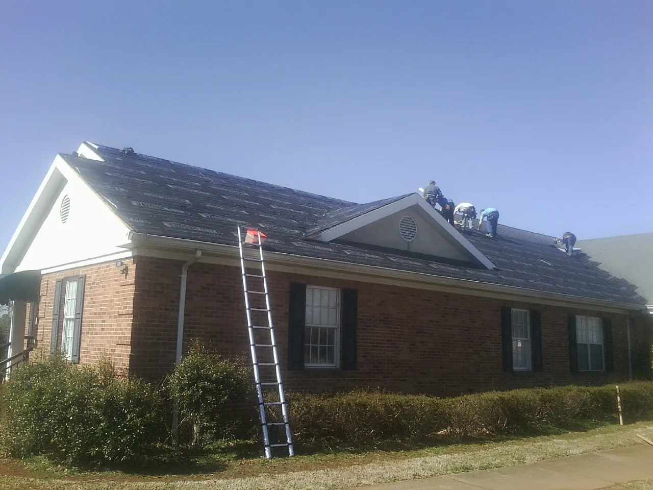 Crew tearing shingles off a brick ranch roof with a ladder set — Emergency Roof Repair work by our crew in Elkins
