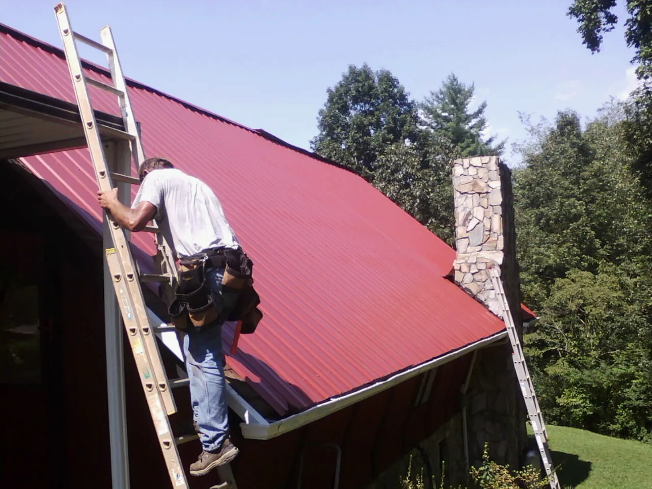 Roofer on a ladder beside a red metal roof and stone chimney — Soffit & Fascia Repair by Roofing Brothers in Elkins